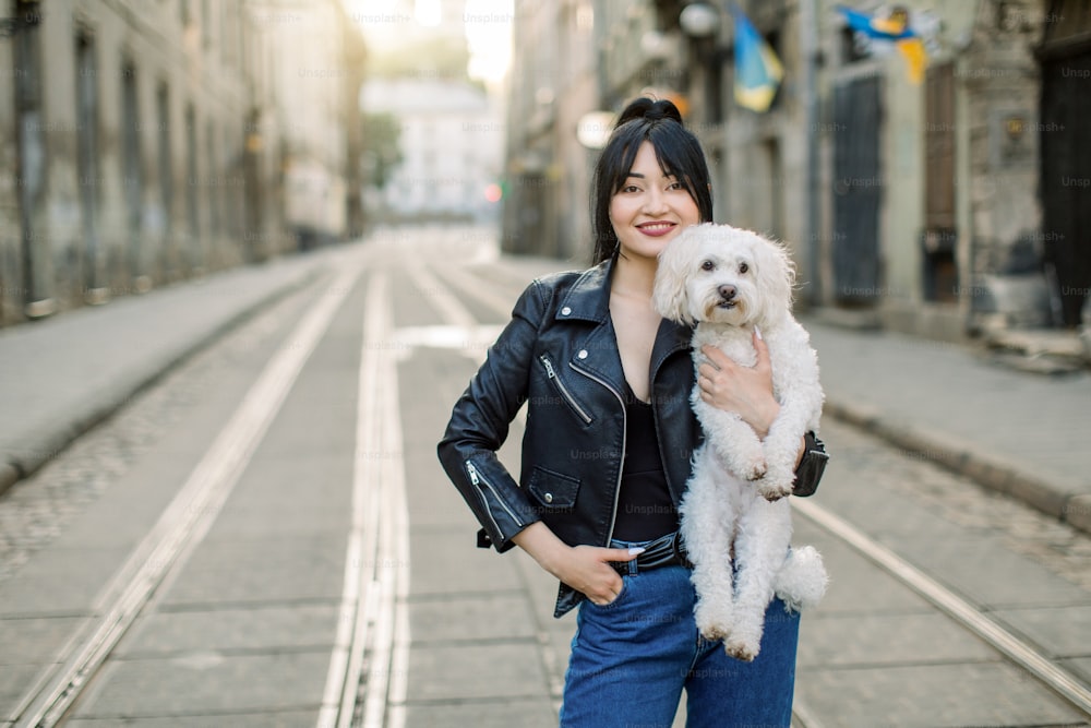 ジーンズコートと黒い革のジャケットを着たファッショナブルなブルネットのアジア人女性が、小さな白い犬を抱いて通りを歩いています。ファッション屋外の都市写真。スペースをコピーします。