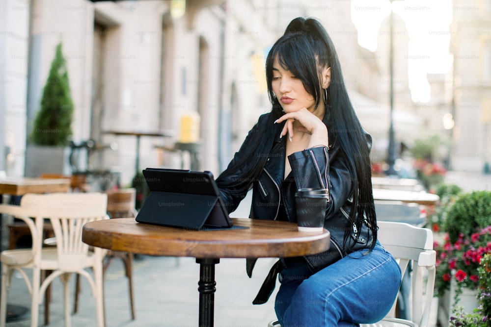 Beautiful concentrated asian young businesswoman, wearing jeans and black leather jacket, sitting at the outdoor city cafe, using digital tablet and drinking coffee. Girl freelancer at cafe.