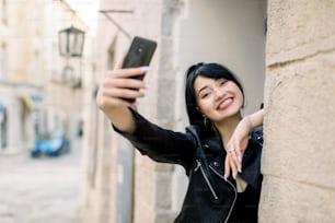 Lifestyle portrait of young happy mixed raced Asian girl, using mobile phone, smiling, making selfie photo and having fun outdoors in the old city street. Morning city