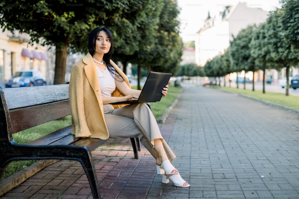 Jovem empresária asiática bastante inteligente concentrada, vestindo elegante terno bege amarelo colorido, sentada no banco da cidade com seu laptop. Wi-Fi gratuito na cidade, conceito de trabalho freelance.