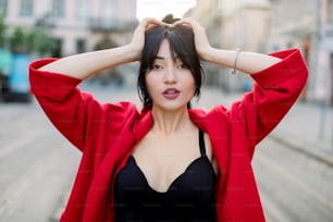 Outdoor portrait of young attractive confident Asian woman, wearing black top and stylish red blazer, posing on the street of old European city, with arms raised to her head. Urban fashion.
