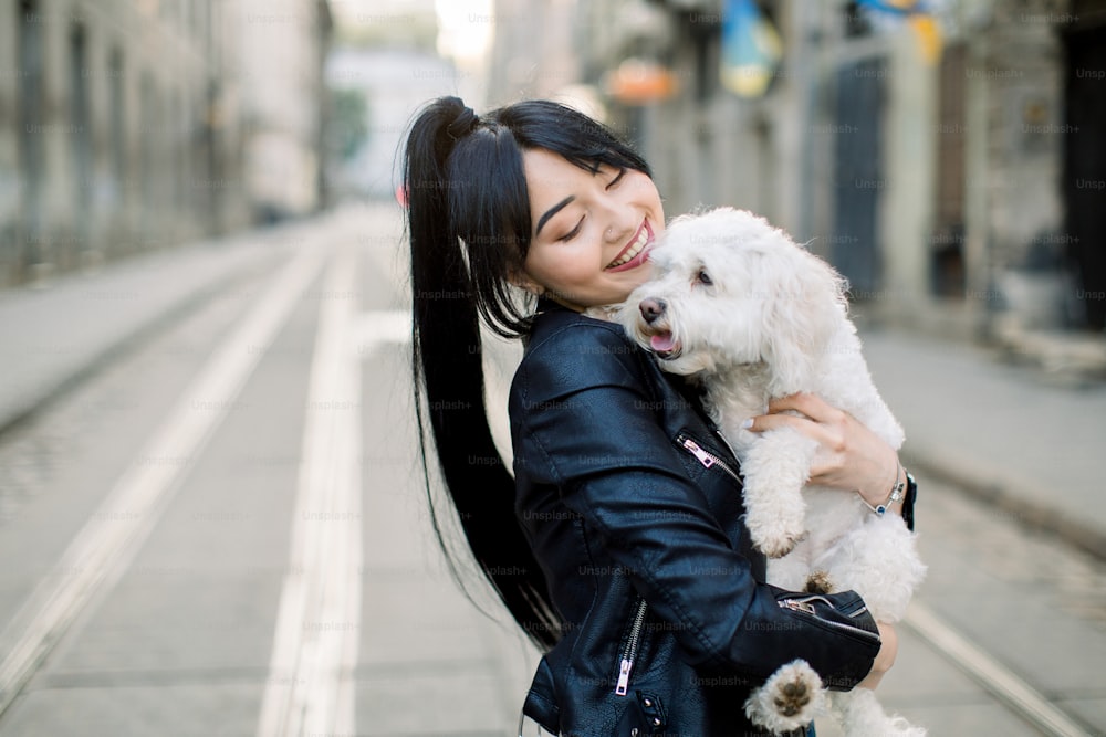 A young dark haired Asian woman, dressed in black leather jacket, stands on street in old European city street, holding and embracing white little dog. Horizontal urban shot, copy space.