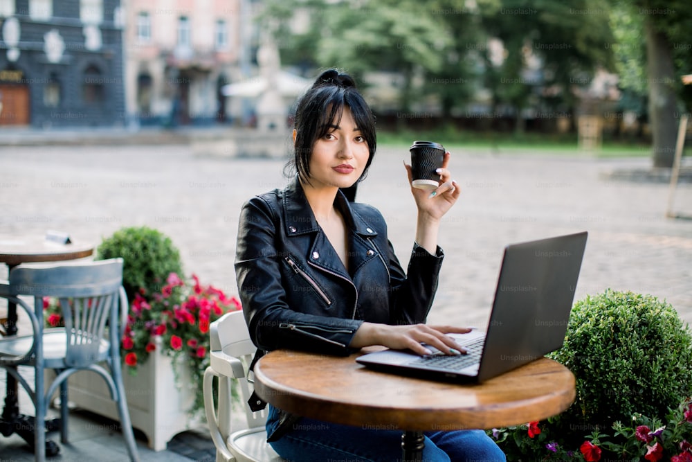 革ジャンを着たアジア系白人女性が、街のカフェのテーブルに座り、ノートパソコンを使いながらコーヒーを飲んでいる。レジャー、仕事、テクノロジーのコンセプト