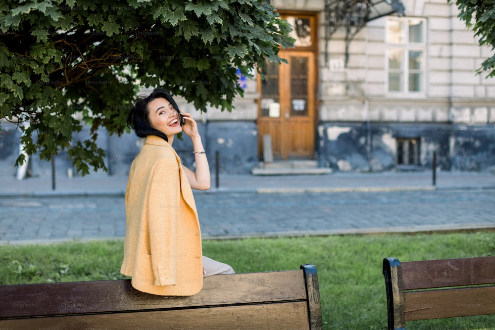 Mujer asiática elegante y risueña con chaqueta amarilla, relajándose y divirtiéndose durante su paseo matutino por la ciudad. Foto al aire libre de encantadora dama de negocios morena sentada en el banco