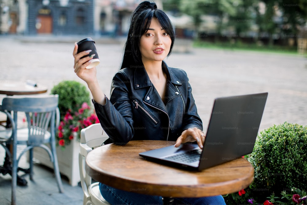 Personas, estilo urbano, concepto de trabajo y ocio. Bonita mujer de negocios asiática con chaqueta de cuero negro, usando una computadora portátil y bebiendo café mientras pasa tiempo en la cafetería
