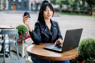 Pessoas, estilo urbano, conceito de trabalho e lazer. Empresária asiática bonita em jaqueta de couro preta, usando laptop e bebendo café enquanto passa o tempo na cafeteria