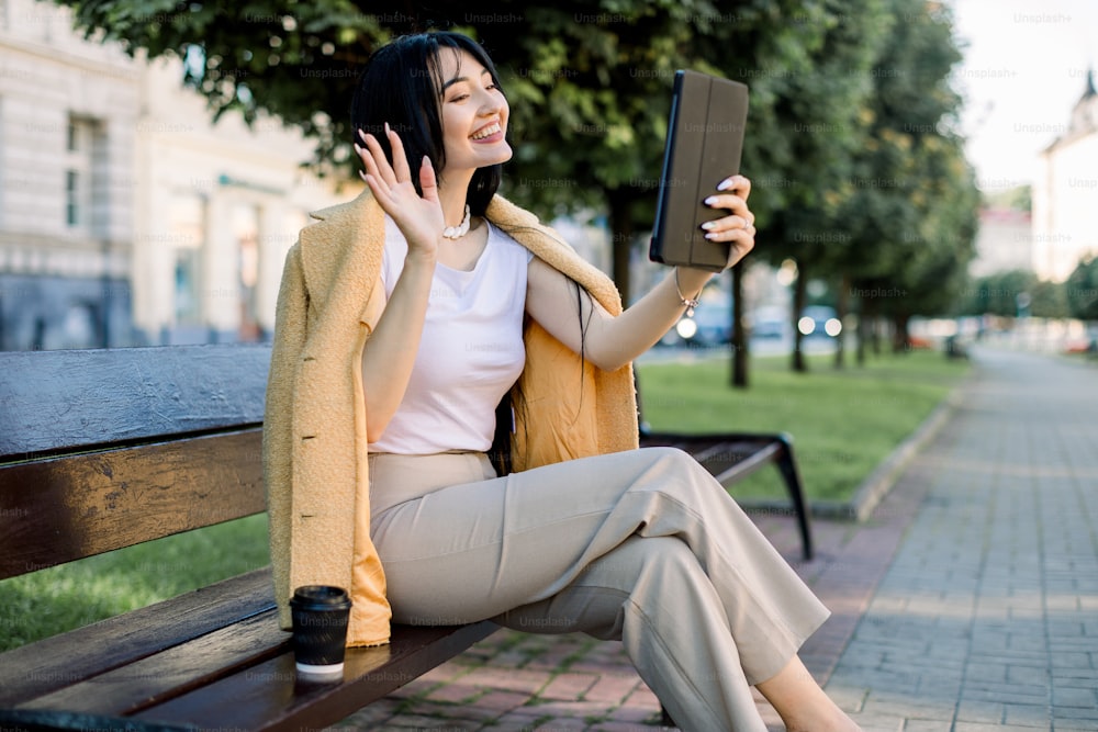 Feliz joven estudiante asiática o empresaria con una tableta y una taza de café desechable, sentada en el banco en un parque de la ciudad de verano y divirtiéndose durante la videollamada, saluda a su amiga.