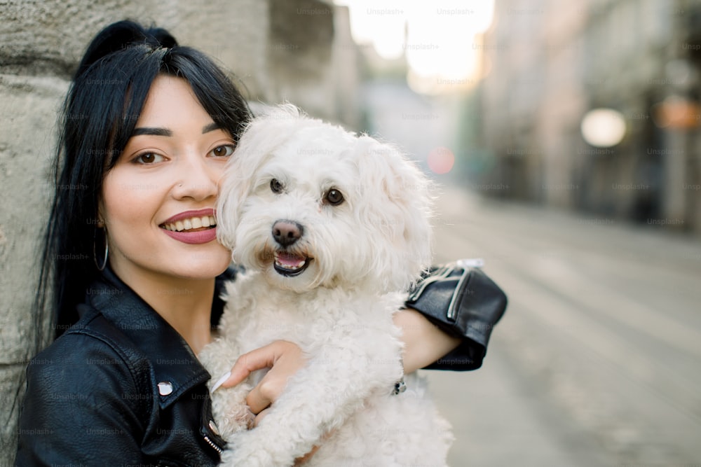 Close up outdoor portrait of lovely young smiling Asian woman in black leather jacket, enjoying the moment, while walking in the city with her little dog. Walk with pet in the city.