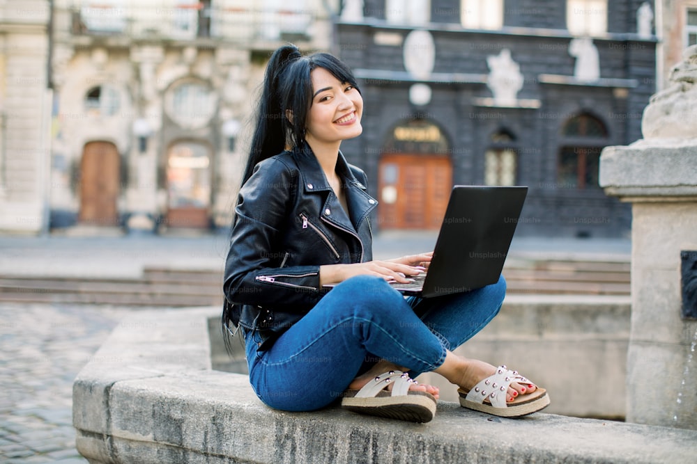 Free Wi-Fi in the city, freelance concept. Smiling young mixed raced Asian woman with black ponytail hair sitting on fountain and texting message on laptop, checking news in social networks