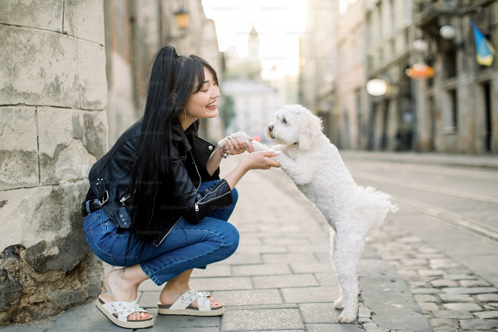 Young Asian urban girl wearing stylish casual clothes, jeans and leather jacket, walks with little cute white dog on the street against the background of the wall, and old vintage buildings.