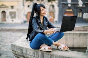 Happy hipster young mixed raced woman working on laptop in the city. Lovely Asian woman with ponytail hair, sitting on vintage stone fountain, studying and using laptop