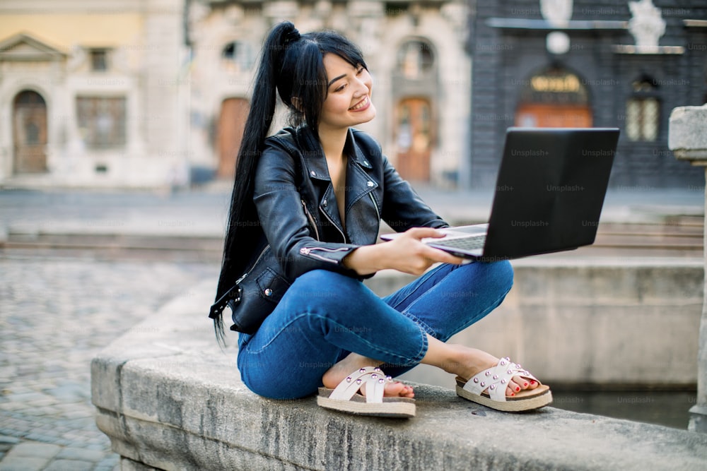 Feliz mujer hipster joven de raza mixta que trabaja en una computadora portátil en la ciudad. Encantadora mujer asiática con pelo de cola de caballo, sentada en una fuente de piedra vintage, estudiando y usando una computadora portátil