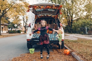 Trick ou tronc. Enfant célébrant la fête traditionnelle d’octobre Halloween dans le coffre de la voiture. Jolie fille heureuse jetant des feuilles dans l’air à l’extérieur. Distanciation sociale et célébration alternative en toute sécurité.