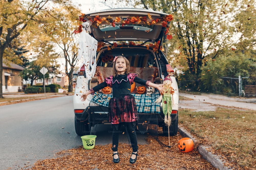 Trick oder Kofferraum. Kleinkind feiert den traditionellen Oktoberfeiertag Halloween im Kofferraum des Autos. Süßes glückliches Mädchen, das Blätter in die Luft wirft. Soziale Distanz und sichere alternative Feier.