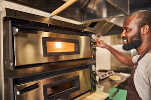 エプロンを着た陽気な若いハンサムな男はオーブンに立っていて、中のピザを見ています
