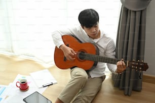 Young man playing a guitar while sitting on floor at home.