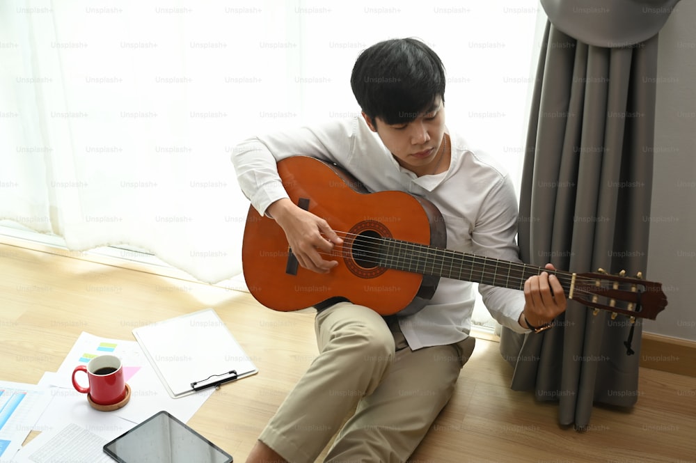 Young man playing a guitar while sitting on floor at home.