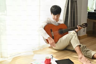 Un hombre joven está sentado en casa junto a la ventana y toca la guitarra acústica en casa.