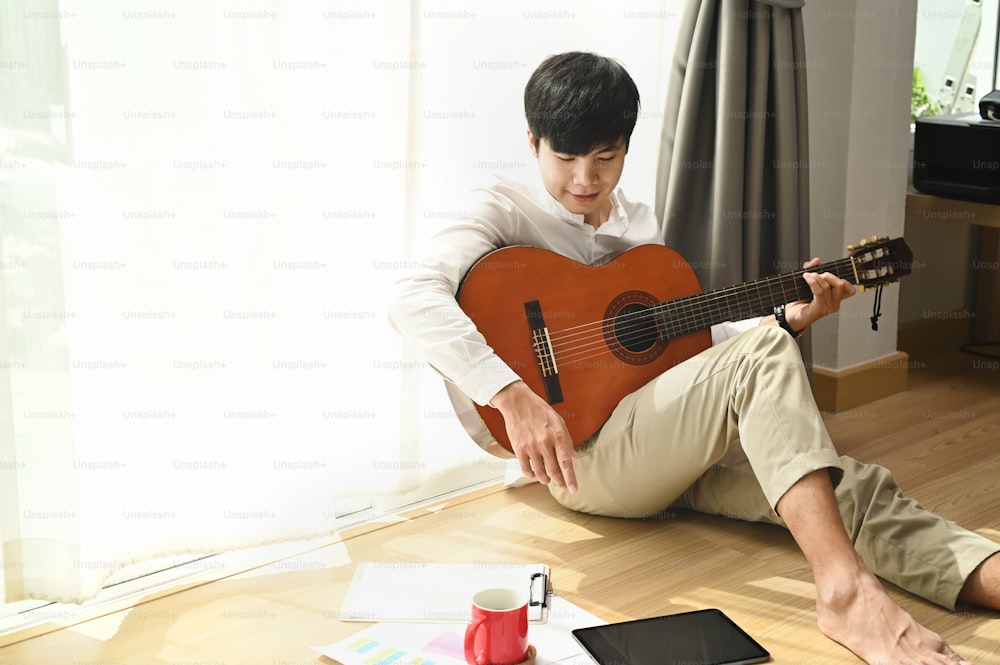 Young male is sitting at home next to the window and playing the acoustic guitar at home.