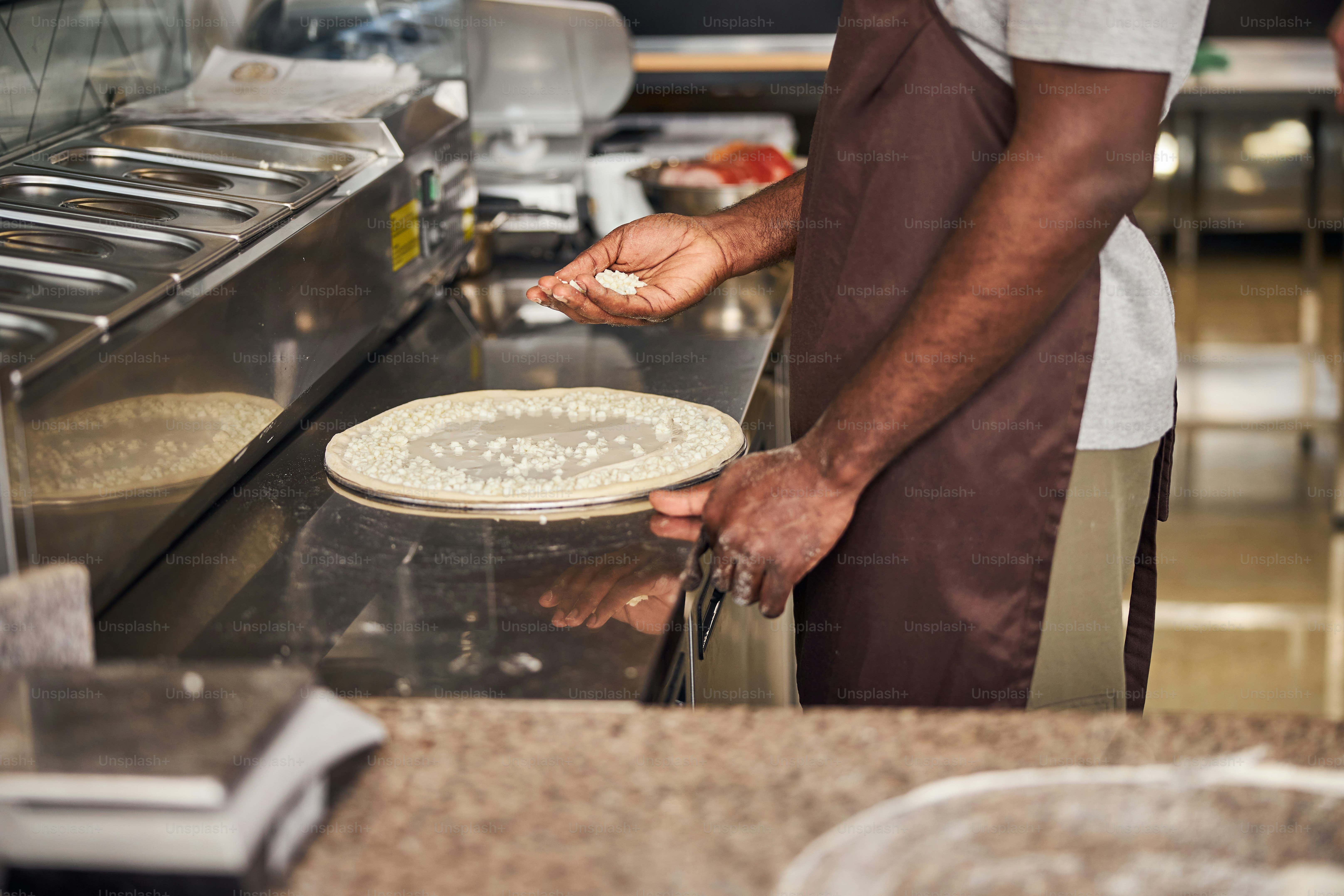 Cropped head of chef in apron standing at table and spreading grated cheese on dough