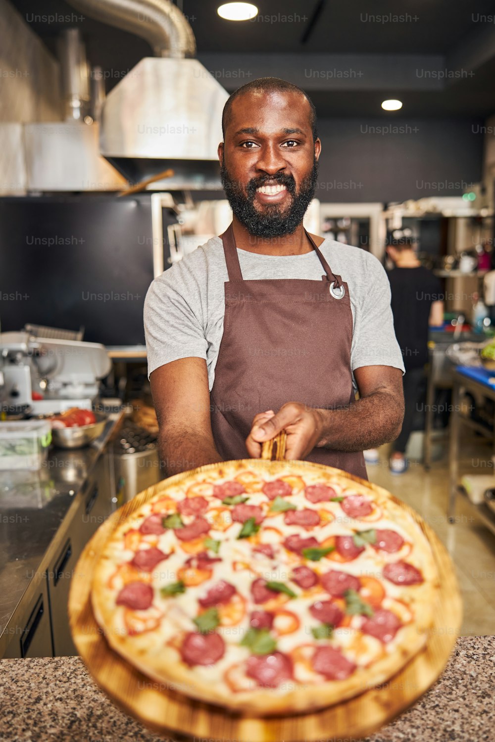 Handsome Afro American man in apron looking at camera and smiling while holding delicious pizza with salami, cheese and tomatoes