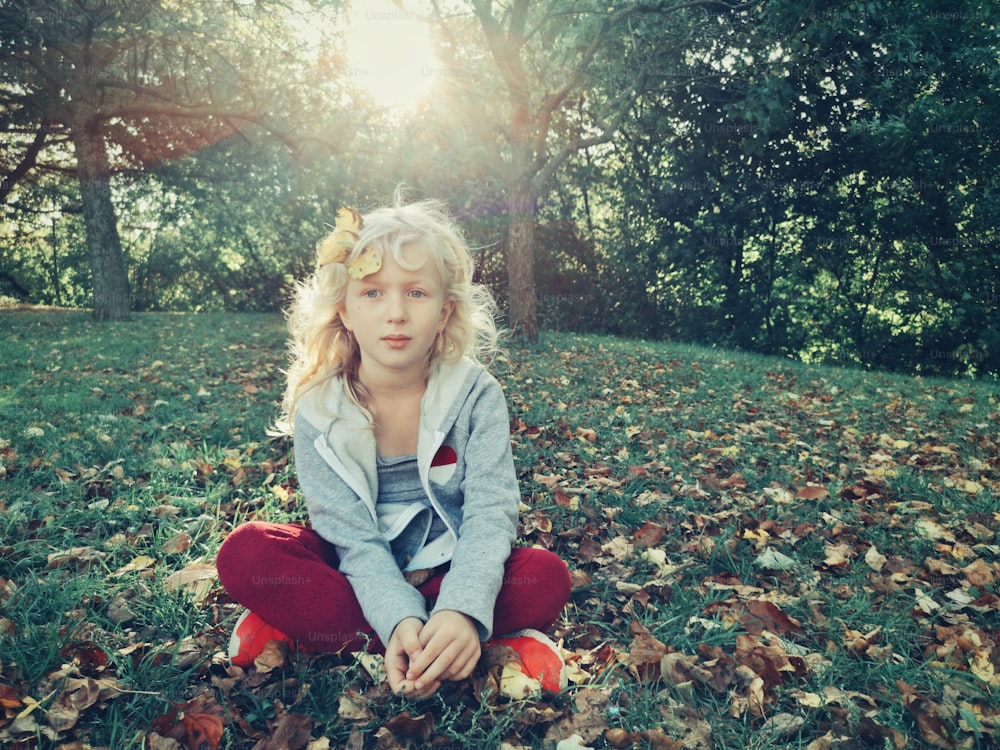 Beautiful pretty preschool Caucasian blonde girl sitting on grass in park outdoor on autumn fall day. Portrait of lovely cute adorable sporty kid outside at sunset.