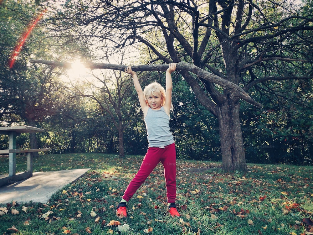 Sporty healthy strong young girl lifting wooden stick branch log in park. Cute kid playing outdoor on summer autumn fall day. Powerful female child girl lifting weights outside.
