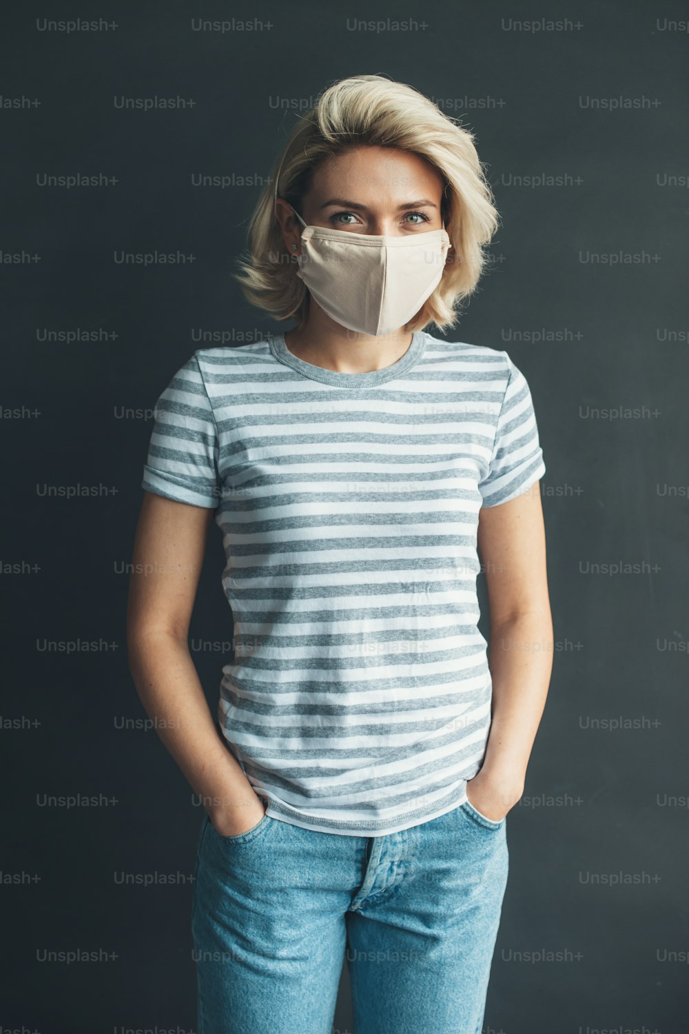 Front view photo of a caucasian woman with medical mask on face posing at camera in a casual clothes on a dark gray studio wall