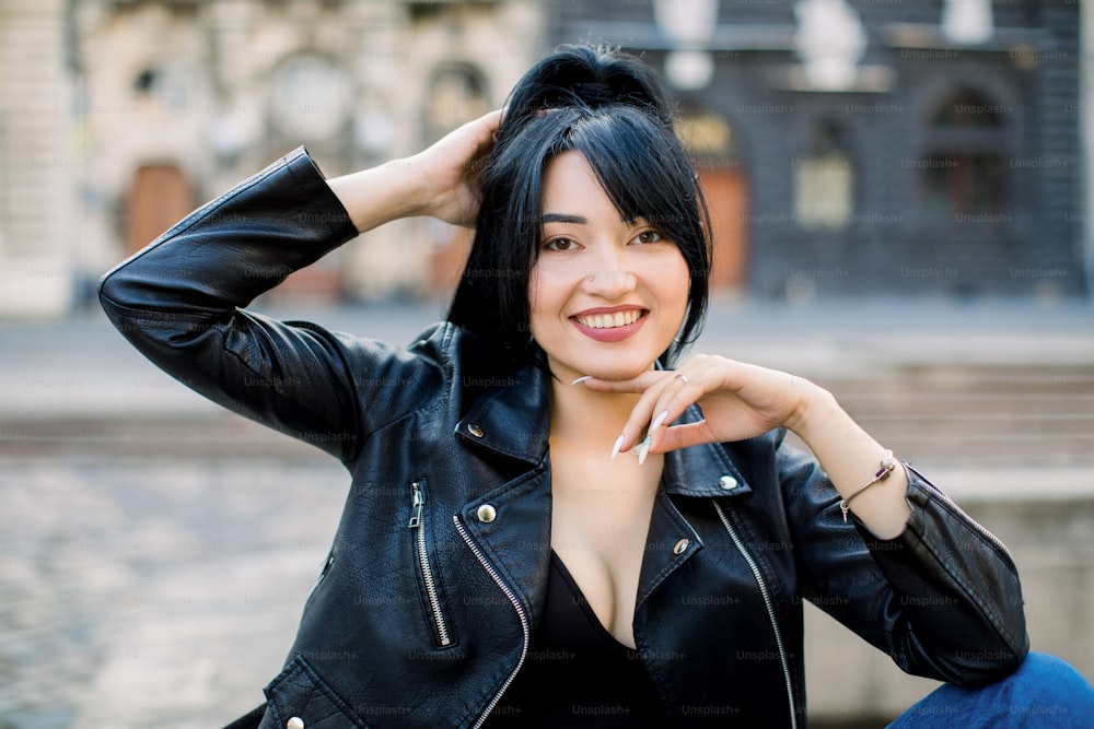 Outdoor close up portrait of young beautiful confident Asian woman, wearing leather jacket, posing on the street of old European city, smiling at camera. Sunny morning. City lifestyle.