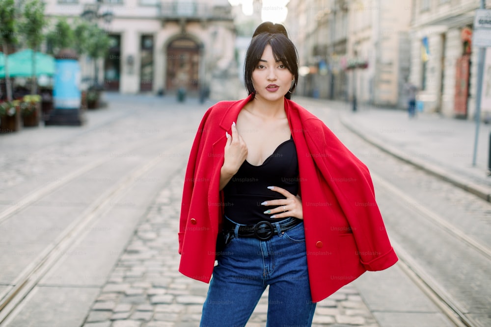 スタイリッシュな赤いジャケット、黒いトップス、ジーンズを着て、黒の長いポニーテールの髪をしたエレガントでかわいいアジアの女性が、夏の日の朝、旧市街の通りでカメラに向かってポーズをとっています。都会のストリートポートレート。