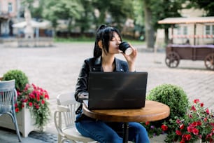 Working online, freelance concept. Young pretty smart Asian woman enjoying the coffee-break, using laptop computer, sitting at the table in city cafe outdoor.