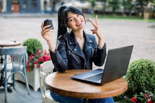 Beautiful smiling cute asian young businesswoman, dressed in black leather jacket, with ponytail hair, sitting at the table in outdoor cafe, using laptop, drinking coffee, and showing victory sign.
