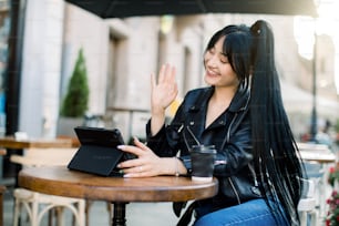 Wi-Fi gratuito, persone, concetto di tecnologie. Bella giovane donna bruna in giacca di pelle, che si diverte durante la videochiamata con l'amico, sorridente e salutante, seduta all'aperto nel caffè della città.