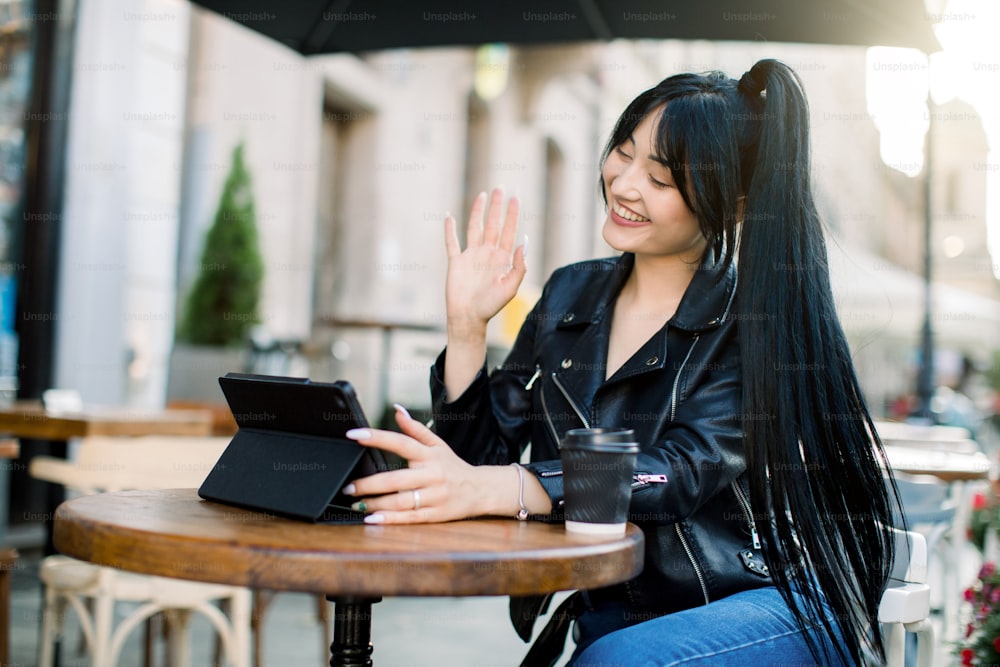Free Wi-Fi, people, technologies concept. Beautiful young brunette woman in leather jacket, having fun during video call with friend, smiling and waving, sitting outdoors in city cafe.