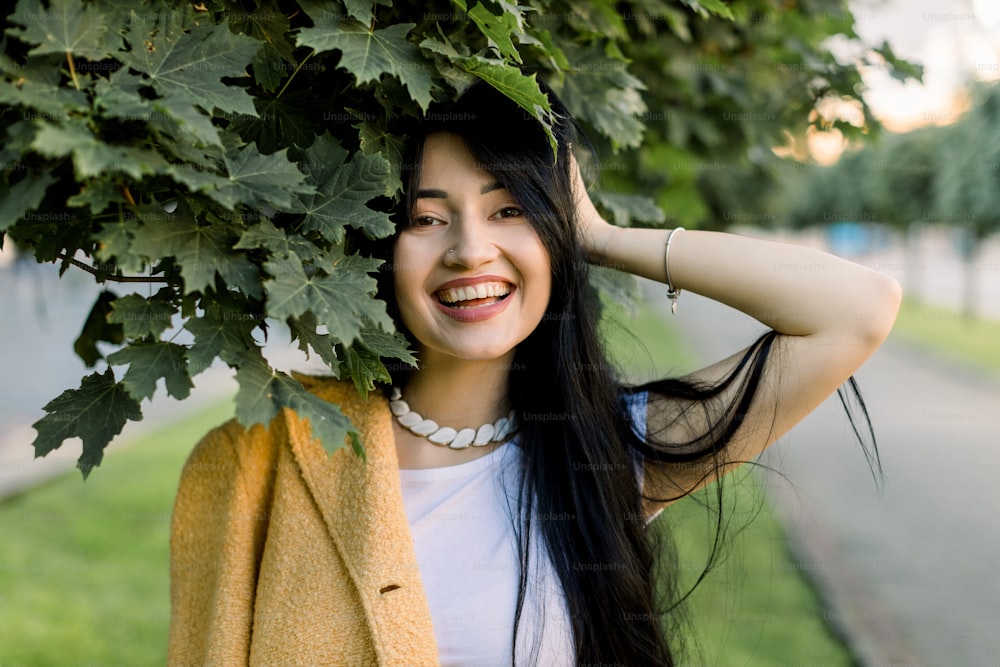 Junges, schönes, fröhliches asiatisches brünettes Mädchen in stilvollem gelbem Blazer und weißem T-Shirt, posiert auf der Stadtstraße unter Ahornbaum und lächelt in die Kamera. Frühlings- und Sommermodekonzept.