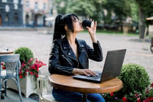 Working online, freelance concept. Young pretty smart Asian woman enjoying the coffee-break, using laptop computer, sitting at the table in city cafe outdoor.