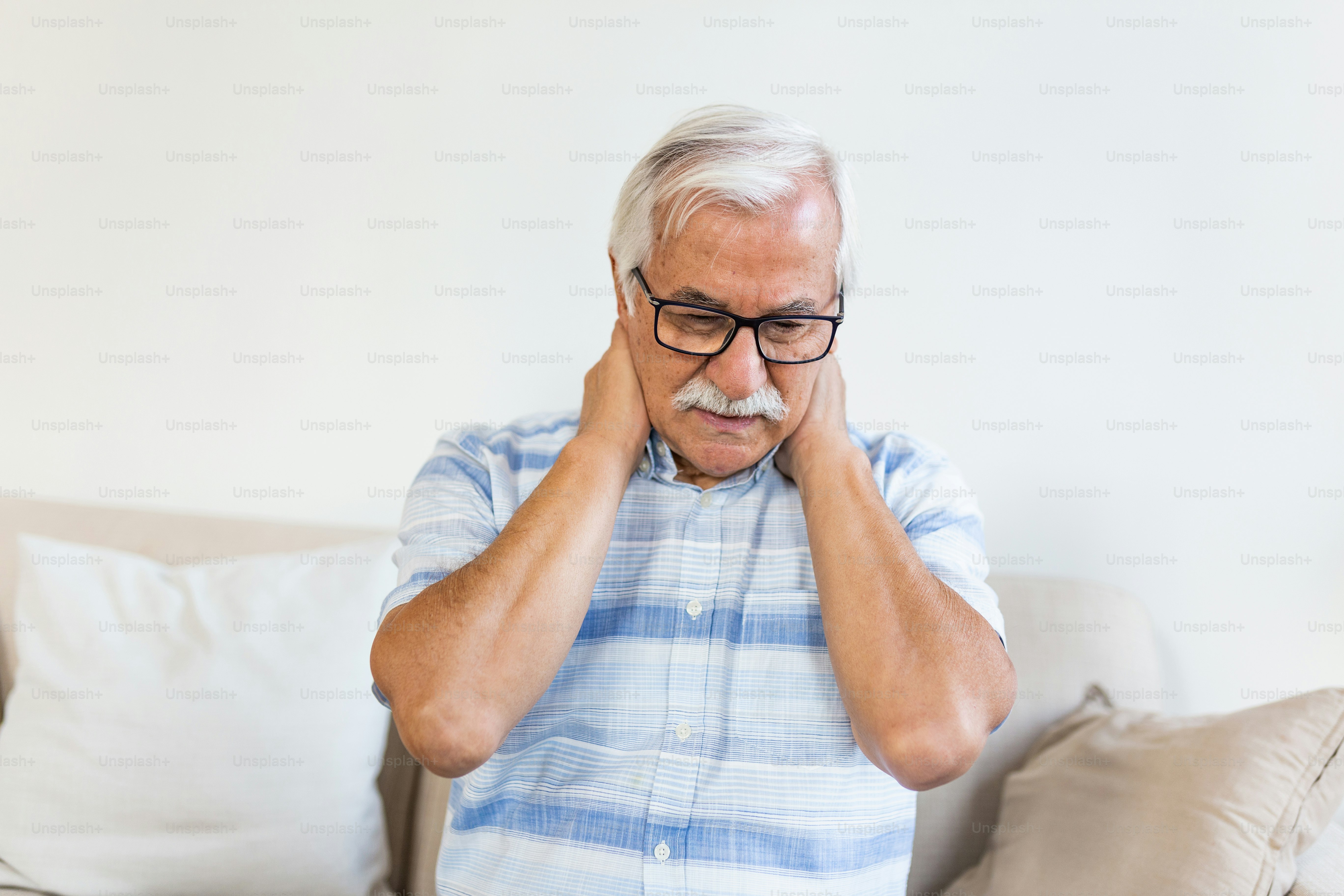 Un homme plus âgé avec des lunettes, se tenant le cou de douleur alors qu’il est assis à l’intérieur