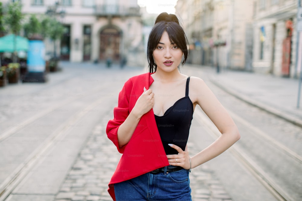 Bella giovane donna sexy asiatica sensuale, che indossa jeans, top nero e blazer rosso alla moda su una spalla, in posa per la macchina fotografica mentre si trova all'aperto nella strada della città vecchia. Stile di vita urbano.