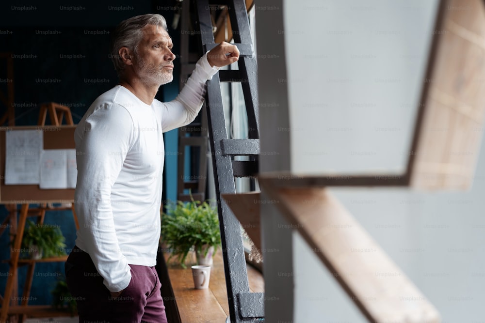 Business lifestyle. Casual Grey-haired Mature professional handsome businessman standing near window in his office while thinking about his goals