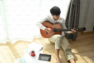 自宅の床に座りながらアコースティックギターを弾く若いアジア人男性。