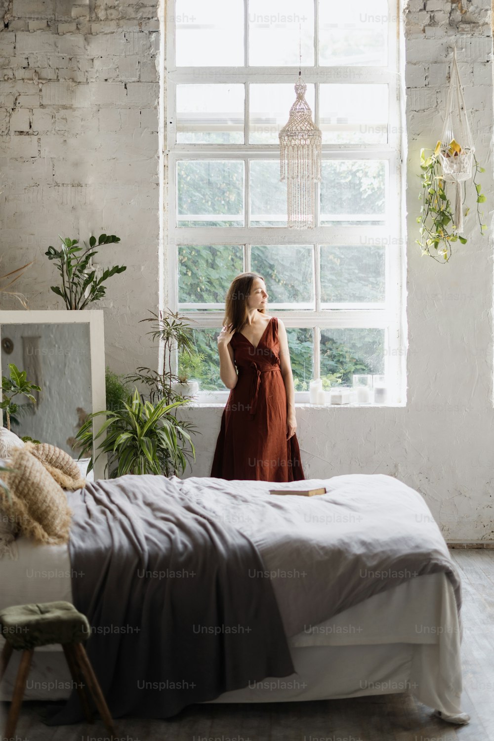 Vertical view of dreamy young woman standing near window at cozy bedroom with interior design in boho chic style. Female enjoying early weekend morning