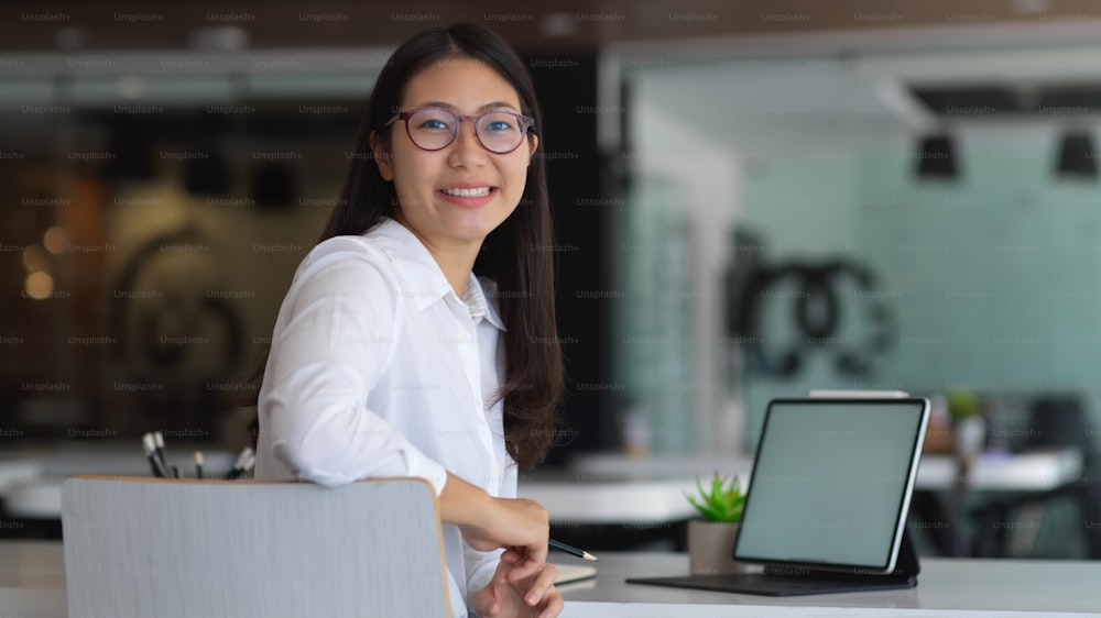 Portrait of female office worker smiling and looking into camera while working with mock up tablet in office