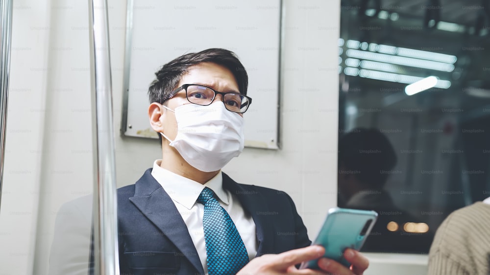 Traveler wearing face mask while using mobile phone on public train . Coronavirus disease or COVID 19 pandemic outbreak and urban city lifestyle problem in rush hour commuting concept .