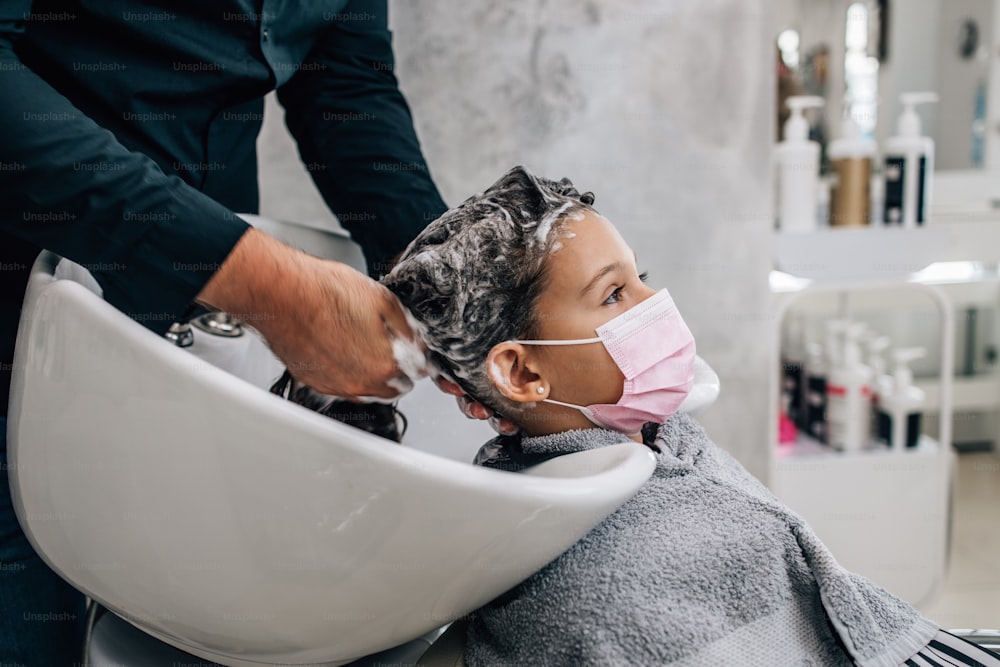 Young girl with protective face mask at hairstyle treatment while professional hairdresser gently washing her hair. Coronavirus lifestyle and new normal concept.