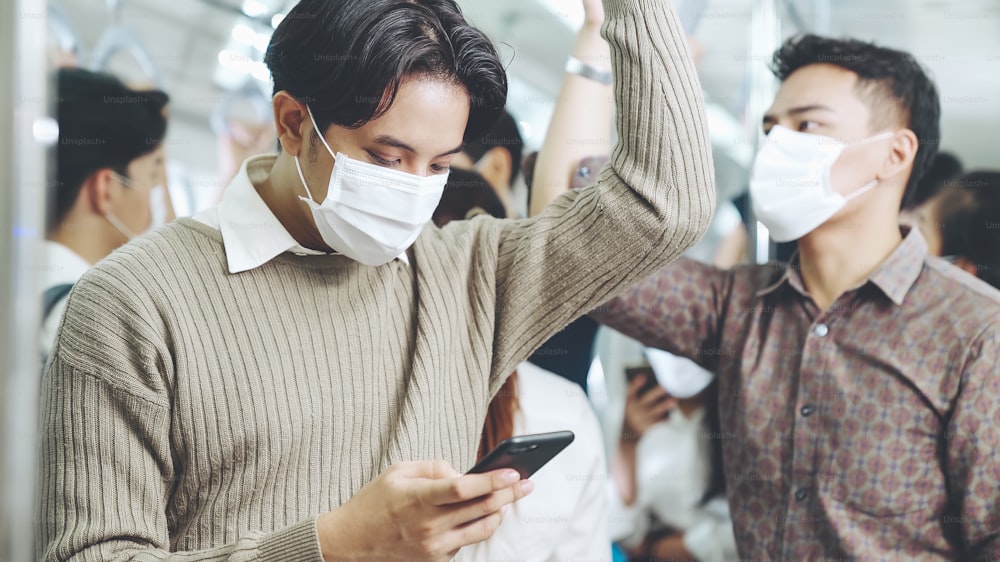 Traveler wearing face mask while using mobile phone on public train . Coronavirus disease or COVID 19 pandemic outbreak and urban city lifestyle problem in rush hour commuting concept .