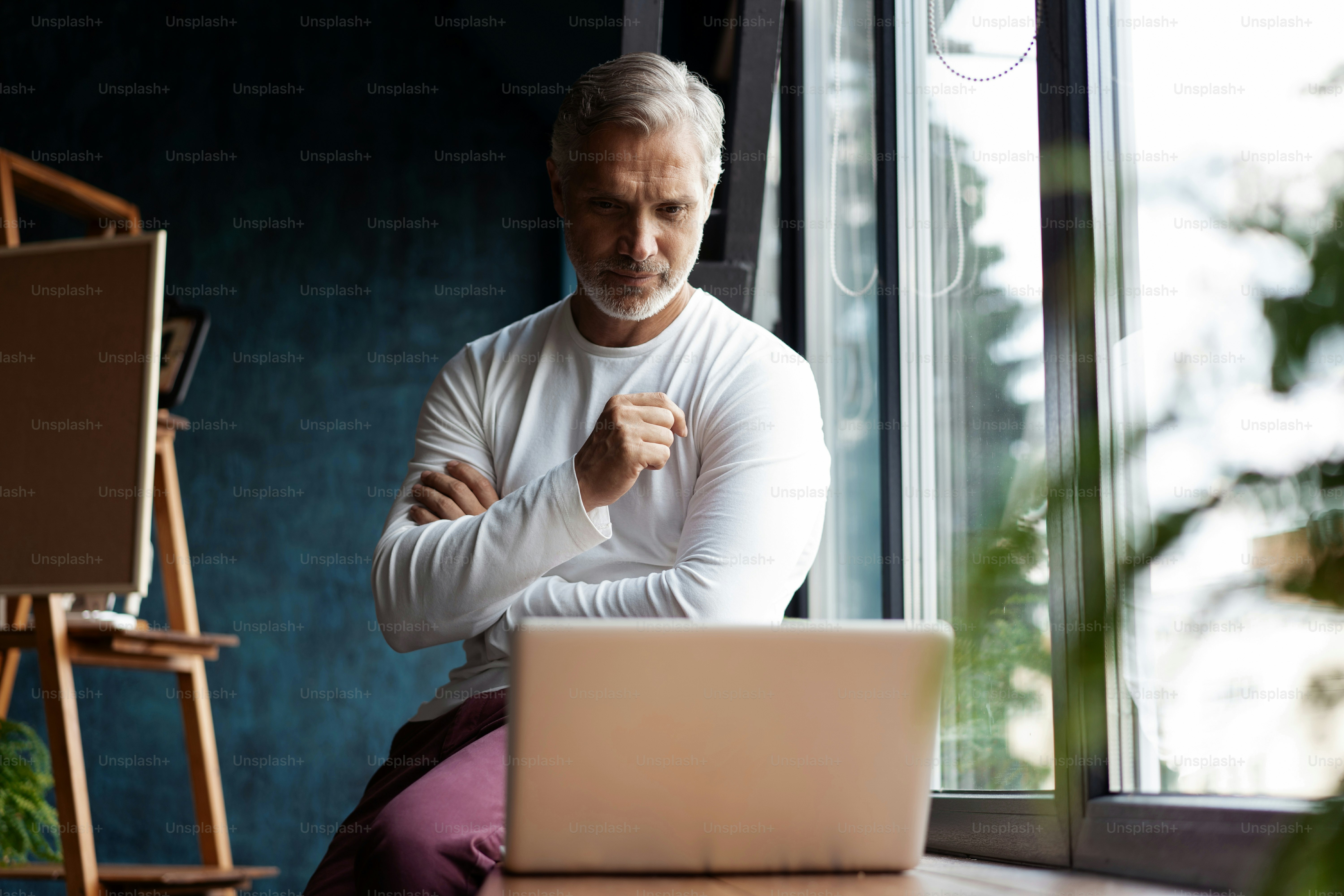 Concentrated on work. Casual Grey-haired Mature man in eyeglasses working on laptop while sitting in office or cafe near the window