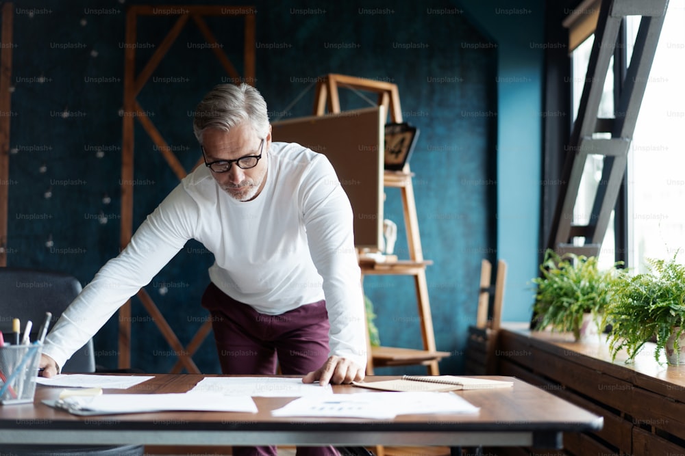 カジュアルな白髪の成熟したハンサムな建築家は、オフィスで建設のスケッチと青写真を見ています