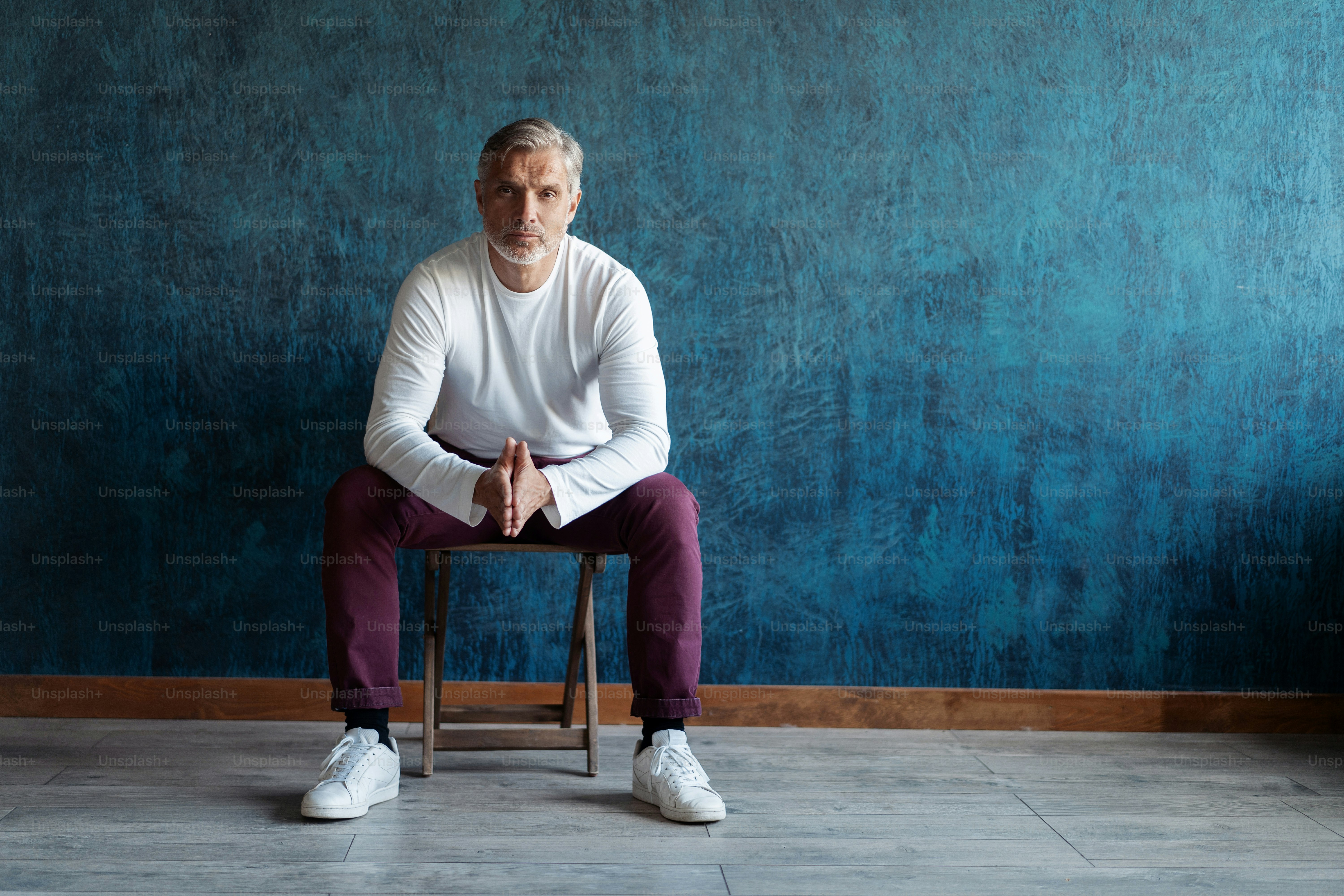 Portrait d’un bel homme mature aux cheveux gris décontracté assis sur une chaise sur fond bleu foncé.