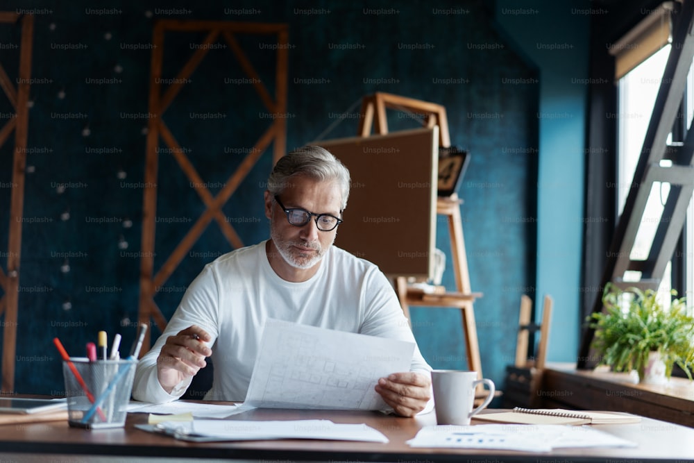 Casual Grey-haired Mature handsome architect looking at blueprint with sketch of construction in office