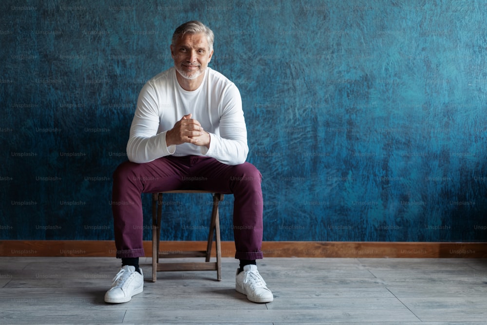Portrait of a casual Grey-haired Mature handsome man sitting on a chair on dark blue background.